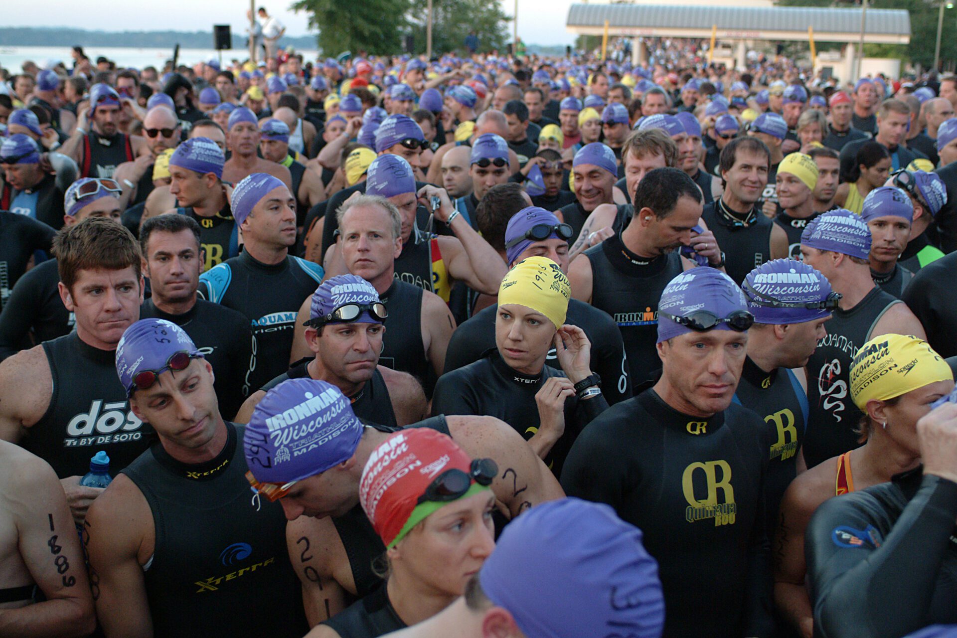Triathletes are shown preparing for the swim portion of the triathlon in wisconsin. They wear purple swim caps and stand together in a crowd ready to max out their training thresholds for the race.