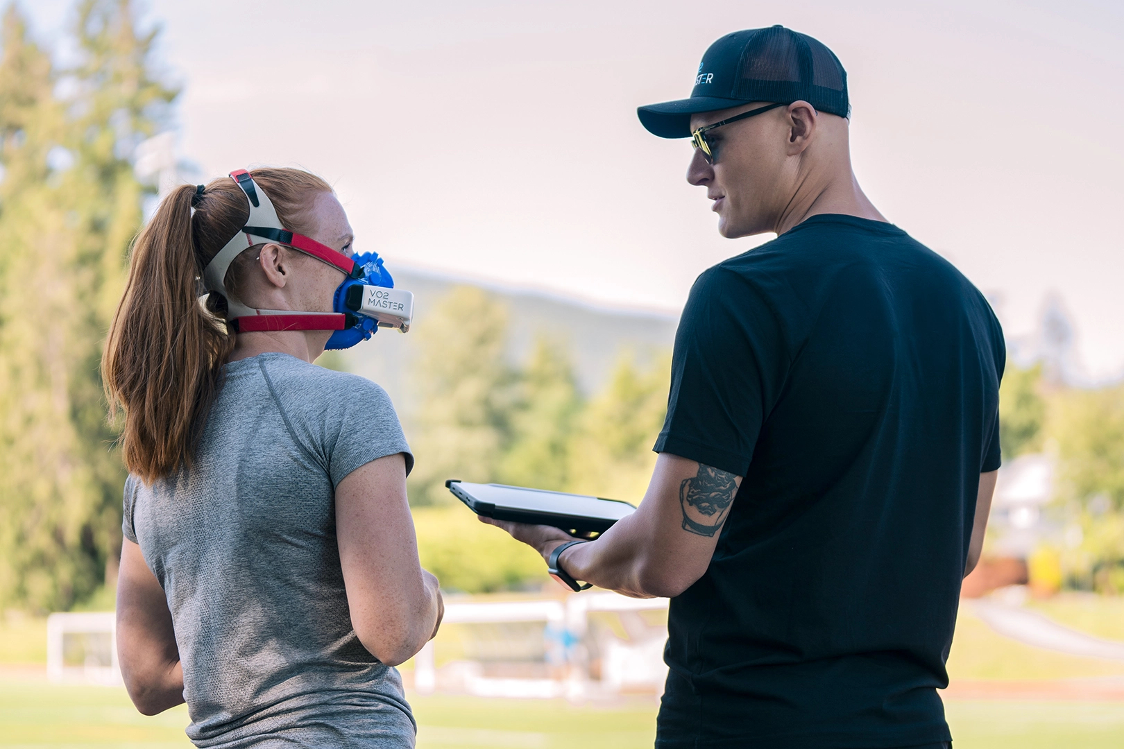 Fitness coach explaining metabolic testing data with a client wearing a portable VO2 analyzer outdoors.